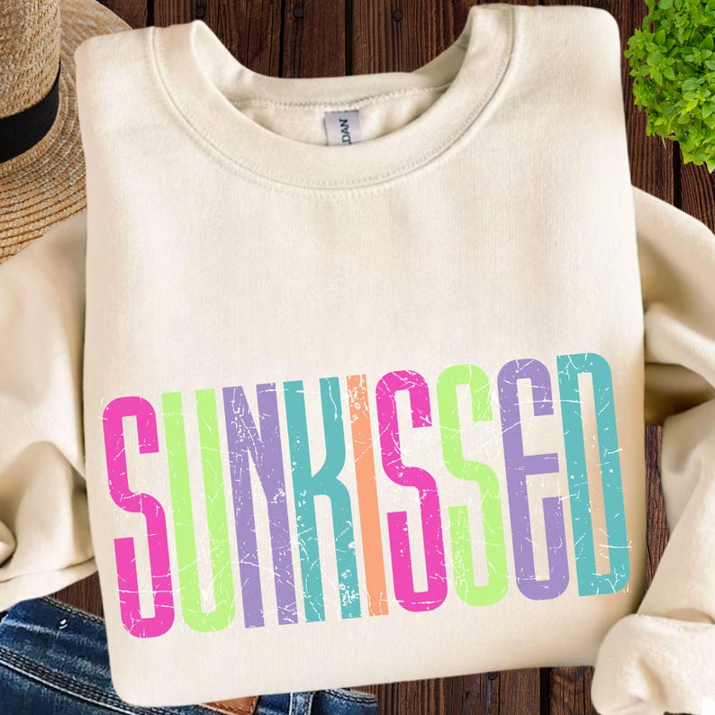 May include: Cream-colored sweatshirt with the word "SUNKISSED" in a colorful, distressed font. The text features pink, green, orange, purple, and blue letters. The sweatshirt is folded on a wooden surface.