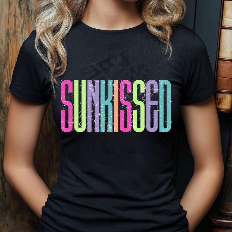 May include: Black t-shirt with the word "SUNKISSED" in large, colorful, distressed lettering. The text features a gradient of pink, yellow, green, and blue. The shirt is a crew neck style.