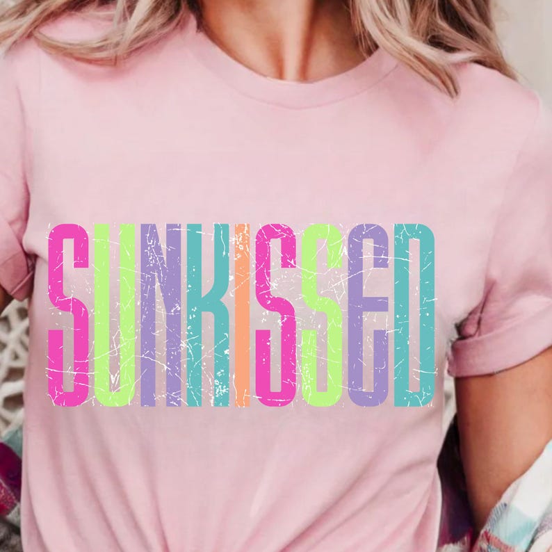 May include: A light pink t-shirt with the word "SUNKISSED" in large, colorful, distressed letters across the front. The letters are in shades of pink, green, orange, and purple. The shirt has short sleeves.