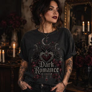 Pode incluir: Uma t-shirt cinzento-carv&atilde;o de manga curta com uma impress&atilde;o gr&aacute;fica de um cora&ccedil;&atilde;o central, rosas e uma lua crescente. O texto "Dark Romance" est&aacute; impresso numa fonte decorativa abaixo do cora&ccedil;&atilde;o com detalhes em vermelho e prata.