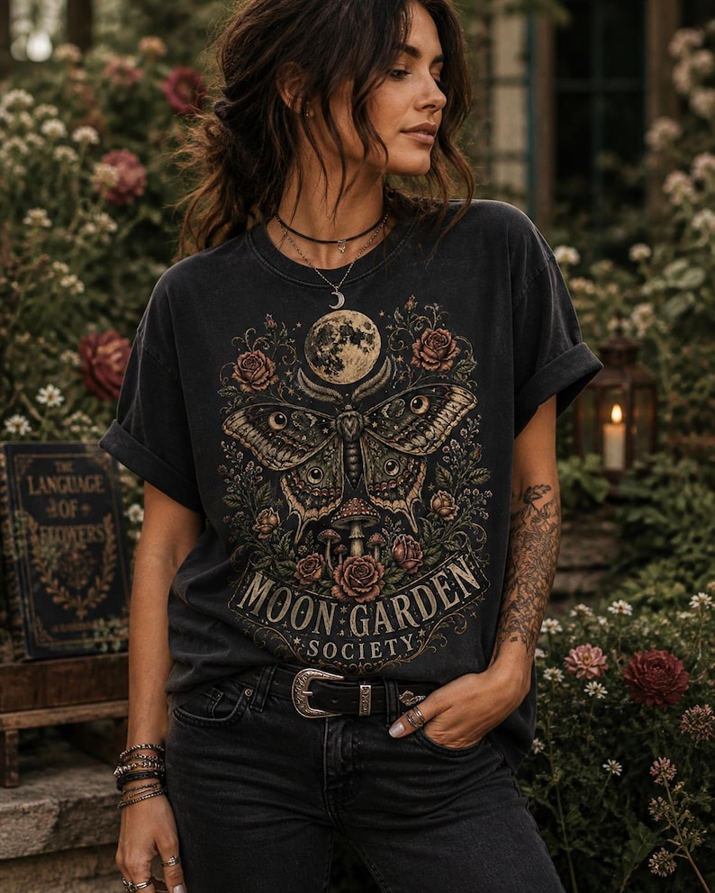 May include: A charcoal gray short-sleeve t-shirt featuring a graphic print of a large moth, a full moon, roses, and mushrooms. The text "MOON GARDEN SOCIETY" is printed in a decorative font at the bottom of the design, which is centered on the chest.