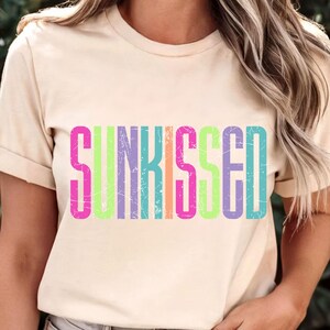 May include: A beige t-shirt with the word "SUNKISSED" printed across the front in bright, colorful letters. The letters are outlined in pink, purple, green, orange, and blue, with a distressed, vintage look.