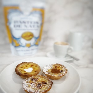 There are 3 portuguese custard tarts in a white plate, one tart is sprinkled with cinnamon, another with powder sugar and the other one with both. There is a cup of coffe in the back and the Baking Kit