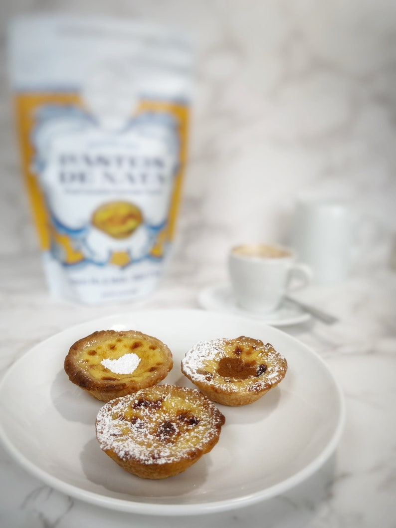 There are 3 portuguese custard tarts in a white plate, one tart is sprinkled with cinnamon, another with powder sugar and the other one with both. There is a cup of coffe in the back and the Baking Kit