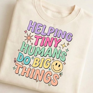 May include: A cream-colored crewneck sweatshirt featuring a multi-colored graphic print that reads HELPING TINY HUMANS DO BIG THINGS in a retro font. The design includes small star shapes and a yellow smiley face icon in pastel colors.