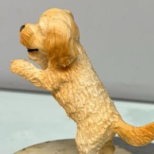 Puede incluir: Una peque&ntilde;a figura de perro de color dorado claro, de pie sobre sus patas traseras con las patas delanteras levantadas. El perro tiene pelaje texturizado, nariz negra y la boca ligeramente abierta, visto de perfil en postura juguetona.