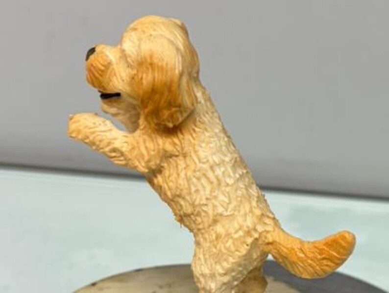 Puede incluir: Una peque&ntilde;a figura de perro de color dorado claro, de pie sobre sus patas traseras con las patas delanteras levantadas. El perro tiene pelaje texturizado, nariz negra y la boca ligeramente abierta, visto de perfil en postura juguetona.