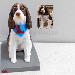 May include: A brown and white Springer Spaniel dog figurine wearing a blue bandana with the text "Ole Miss" in red. The figurine is sitting on a gray base.