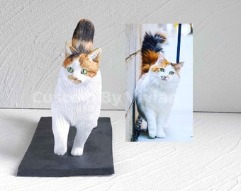 May include: A white, orange, and black cat figurine with green eyes. The figurine is standing on a black base. The cat is looking to the right. A photo of a similar cat is in the background.