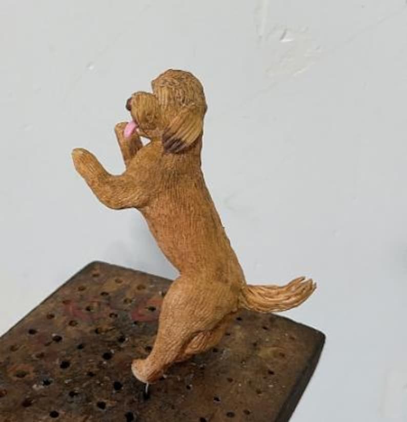 Puede incluir: Una peque&ntilde;a figura de perro de color marr&oacute;n claro de pie sobre sus patas traseras con las patas delanteras levantadas. El perro tiene pelaje texturizado, orejas ca&iacute;das y una peque&ntilde;a lengua rosa visible. Est&aacute; montado sobre una base oscura.