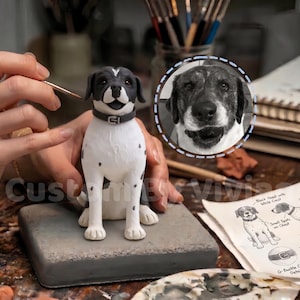 May include: A handcrafted dog figurine with a black head, ears, and spots on a white body. The dog wears a gray collar. A detailed sketch and a portrait of a dog are in the background. The figurine is on a gray base.