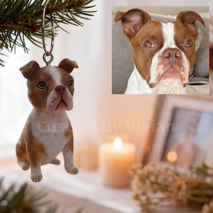 May include: A brown and white dog ornament suspended from a fir tree branch. The ornament is a detailed figurine of a dog with a white chest and face, and brown ears and head. A photo of a similar dog is in the background.