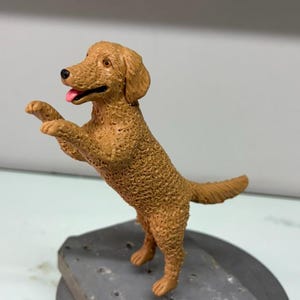 Puede incluir: Una figura de perro de color marr&oacute;n dorado con pelaje texturizado, de pie sobre sus patas traseras con las patas delanteras levantadas. El perro tiene nariz negra, ojos marrones y la boca abierta mostrando una lengua rosa. Base gris.