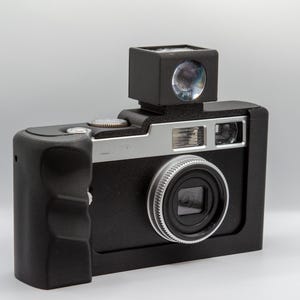 Könnte beinhalten: Eine schwarze und silberne Vintage-Kamera mit einem prominenten Objektiv und Sucher. Die Kamera hat einen rechteckigen Körper mit einem Griff an der Seite. Das Objektiv ist von einem silbernen Ring umgeben. Der Sucher ist eine kleine schwarze Box oben.