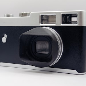 May include: A black and silver digital camera with a lens hood. The camera has a silver top and a black body. The lens is centered on the front of the camera. The camera is on a white surface.