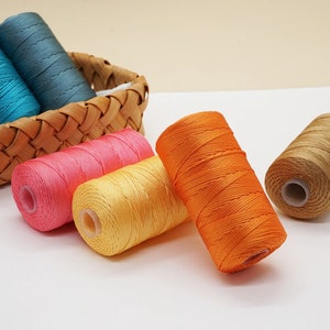 Könnte beinhalten: Vier Spulen mit Stickgarn in verschiedenen Farben: Orange, Gelb, Rosa und Braun. Die Spulen liegen auf einer weißen Oberfläche.