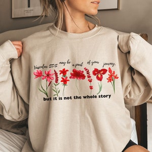 May include: A beige sweatshirt with a floral design and the text "Vascular EDS may be a part of your journey, but it is not the whole story."