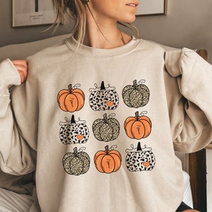 May include: A beige sweatshirt with a graphic of nine orange pumpkins with black and brown leopard print accents. The pumpkins have small hearts on them and some say "Mama" on them.