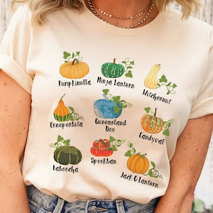 May include: A beige t-shirt with a graphic of different types of gourds and pumpkins with their names written below each one. The gourds and pumpkins include: Pumpkinella, Ninja Lantern, Witchernut, Creeperbita, Queensland Boo, Candyval, Spookban, Kaboocha, and Jack-O-Lantern.