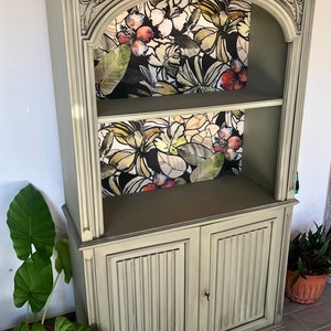 Op de afbeelding: Een hoge, groene houten boekenkast met twee planken en twee deuren. De planken zijn bekleed met tropisch bloemmotief behang. De boekenkast staat op een houten platform met wielen.