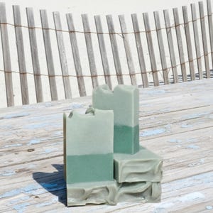 May include: Three rectangular, two-tone green soaps are stacked on a weathered wooden surface. The soaps have a light green top and a darker green bottom. The background features a beach scene with a wooden fence and the ocean.
