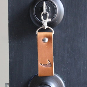 May include: A brown leather keychain with a silver clasp and a metal rivet. The keychain features a cutout of a coastal outline. The keychain is attached to a black door lock.