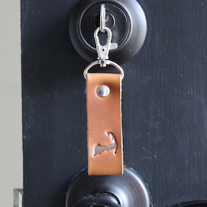 Puede incluir: Un llavero de cuero marrón claro con un mosquetón y un anillo plateados está sujeto a una cerradura de puerta. El llavero presenta un contorno costero. El cuero es de color marrón claro.