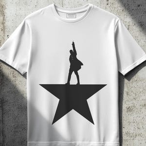 May include: White t-shirt with a black silhouette of a person standing on a star. The person is in a pose with their arm raised and their leg extended. The star is a five-pointed star.