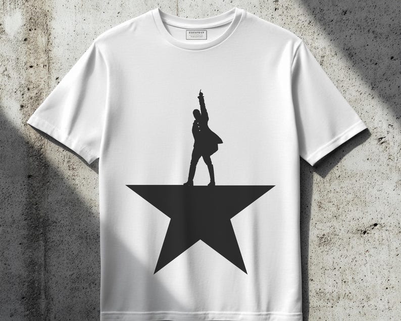 May include: White t-shirt with a black silhouette of a person standing on a star. The person is in a pose with their arm raised and their leg extended. The star is a five-pointed star.