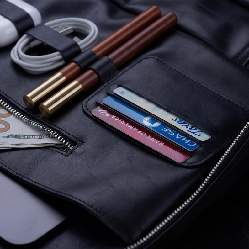 May include: A black leather organizer featuring a zippered compartment containing US currency, a white charging cable, two brown pens with gold-tone caps, and a card slot holding a Bank of America debit card and a Chase Texas debit card.
