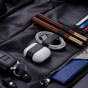 May include: A black leather organizer featuring a white wireless earbud case, a coiled white charging cable, two brown wooden pens with gold accents, a black car key fob, and a blue card holder with a visible portion of a US dollar bill.
