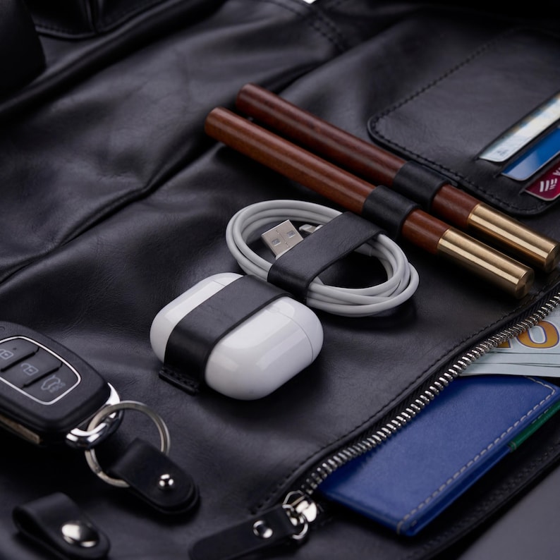 May include: A black leather organizer featuring a white wireless earbud case, a coiled white charging cable, two brown wooden pens with gold accents, a black car key fob, and a blue card holder with a visible portion of a US dollar bill.
