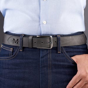 May include: A dark gray leather belt featuring a silver-toned metal buckle and a personalized engraving on the strap. The engraving displays a large letter M with the name Matthew written horizontally across the center of the letter.