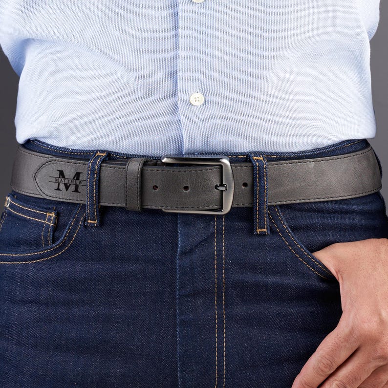 May include: A dark gray leather belt featuring a silver-toned metal buckle and a personalized engraving on the strap. The engraving displays a large letter M with the name Matthew written horizontally across the center of the letter.