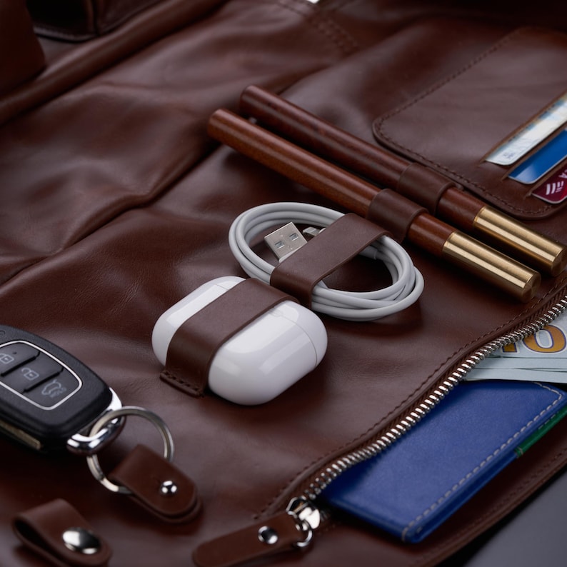 May include: A brown leather organizer featuring a white wireless earbud case, a coiled white charging cable, two brown pens with gold accents, a black car key fob, and a blue card holder with a visible one hundred dollar bill inside the pocket.