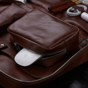 May include: A brown leather backpack featuring a front zippered pocket that contains a white computer mouse. The backpack has silver-toned metal zippers and additional compartments holding a white charging cable and a white power adapter.