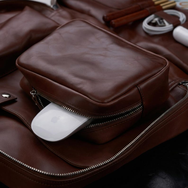 May include: A brown leather backpack featuring a front zippered pocket that contains a white computer mouse. The backpack has silver-toned metal zippers and additional compartments holding a white charging cable and a white power adapter.