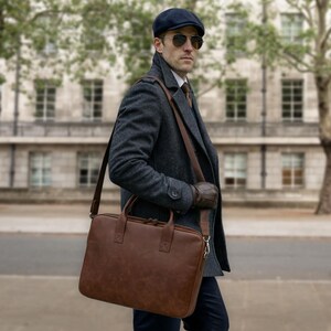 May include: A brown leather briefcase with two top handles and a detachable shoulder strap. The rectangular bag features a zippered main compartment, visible stitching details, and metal hardware attachments for the strap, presented in a solid brown finish.