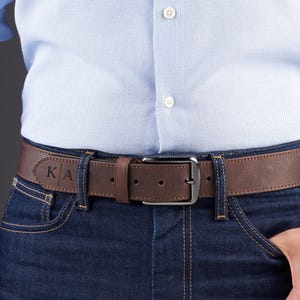 May include: A brown leather belt with a dark metal buckle, worn through the loops of dark blue denim jeans. The belt features a personalized engraving of the letters K and A separated by a vertical line on the left side of the strap.