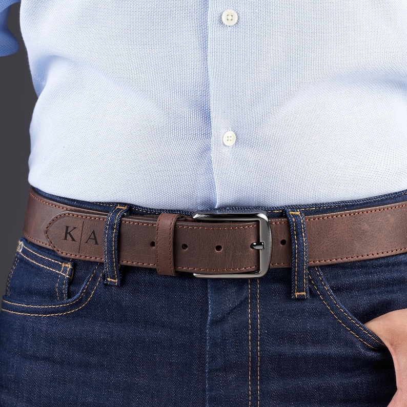 May include: A brown leather belt with a dark metal buckle, worn through the loops of dark blue denim jeans. The belt features a personalized engraving of the letters K and A separated by a vertical line on the left side of the strap.