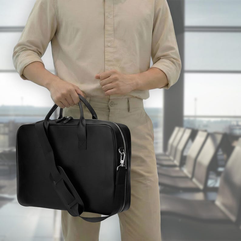 May include: A black rectangular briefcase with a smooth finish, featuring two top handles, a detachable shoulder strap with a padded section, and a silver-toned metal zipper closure that extends along the top and sides of the bag.