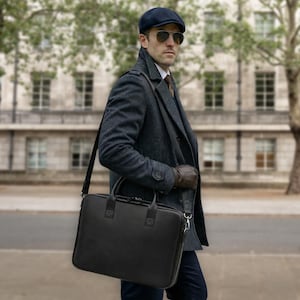 May include: A black rectangular briefcase with a smooth finish, featuring dual top handles and a detachable shoulder strap. The bag has a zippered main compartment and is constructed from a solid material, designed for carrying documents or a laptop.
