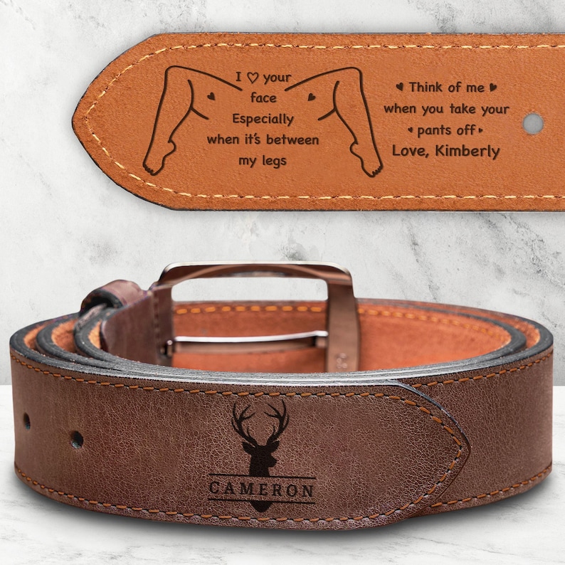 May include: A brown leather belt featuring a silver-toned buckle and a dark brown deer silhouette logo with the name CAMERON engraved below. The inner side displays the text: I heart your face Especially when it's between my legs. Think of me when you take your pants off. Love, Kimberly.