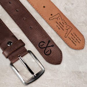 May include: A dark brown leather belt with a silver-toned metal buckle. The inner side of the belt features a black engraved graphic of legs and text that reads: I love your face, especially when it's between my legs. Think of me when you take your pants off. Love, Kimberly.