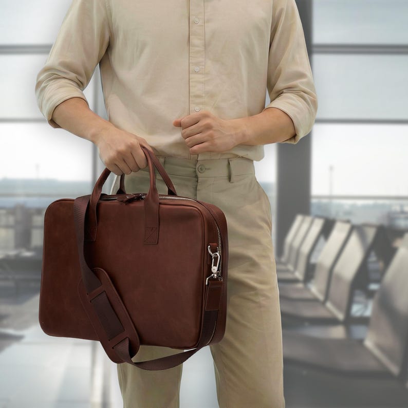 May include: A dark brown leather briefcase featuring two top handles and a detachable shoulder strap with a padded section. The rectangular bag has a zippered main compartment, silver-toned metal hardware, and a smooth, matte finish.