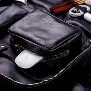 May include: A black leather bag featuring a rectangular front pocket with a silver zipper. A white computer mouse is partially inserted into the open zipper compartment. The bag has a smooth texture and visible stitching along the edges.