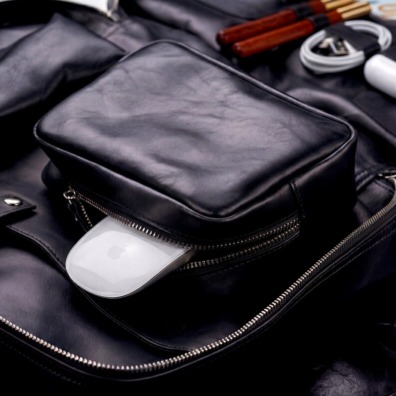 May include: A black leather bag featuring a rectangular front pocket with a silver zipper. A white computer mouse is partially inserted into the open zipper compartment. The bag has a smooth texture and visible stitching along the edges.