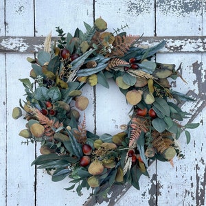 May include: A seasonal wreath featuring artificial leaves in shades of green and brown, with acorns and fern accents. The wreath is displayed on a distressed, white-painted wooden door.