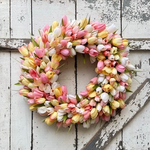 May include: A circular wreath made of artificial tulips in pink, yellow, and white. The wreath is densely packed, giving a full appearance. Green leaves are placed among the flowers. The wreath is set against a weathered, white-painted wooden background.
