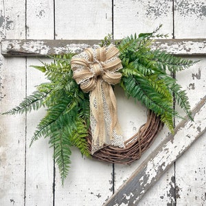 May include: A rustic wreath with a natural grapevine base, decorated with vibrant green fern leaves. A large burlap bow with lace detailing provides a charming accent. The wreath is set against a distressed white wooden backdrop.